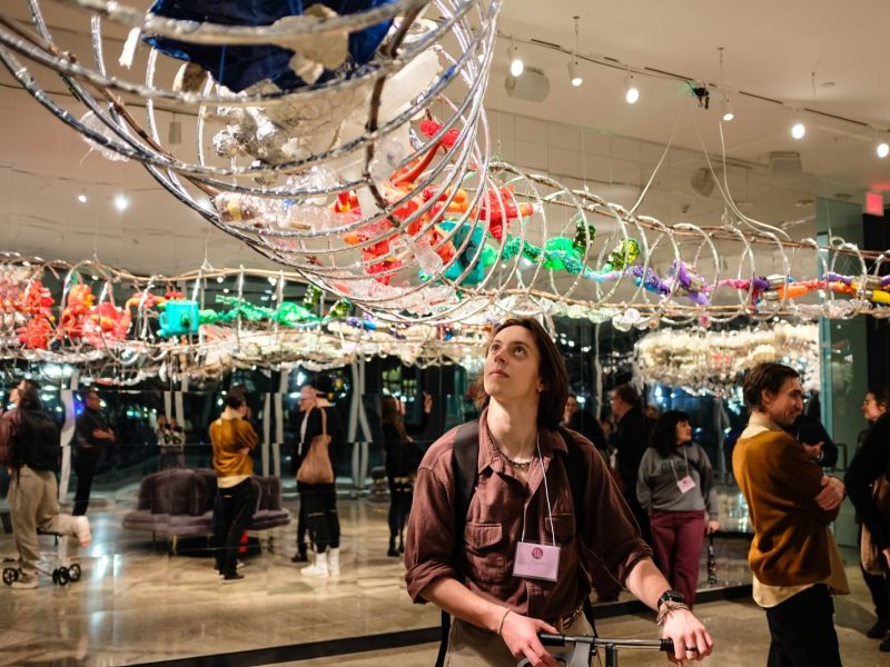 A participant in the University of Michigan Museum of Art's Queer Night stands under artist Machine Dazzle's "Ouroboros" sculpture.