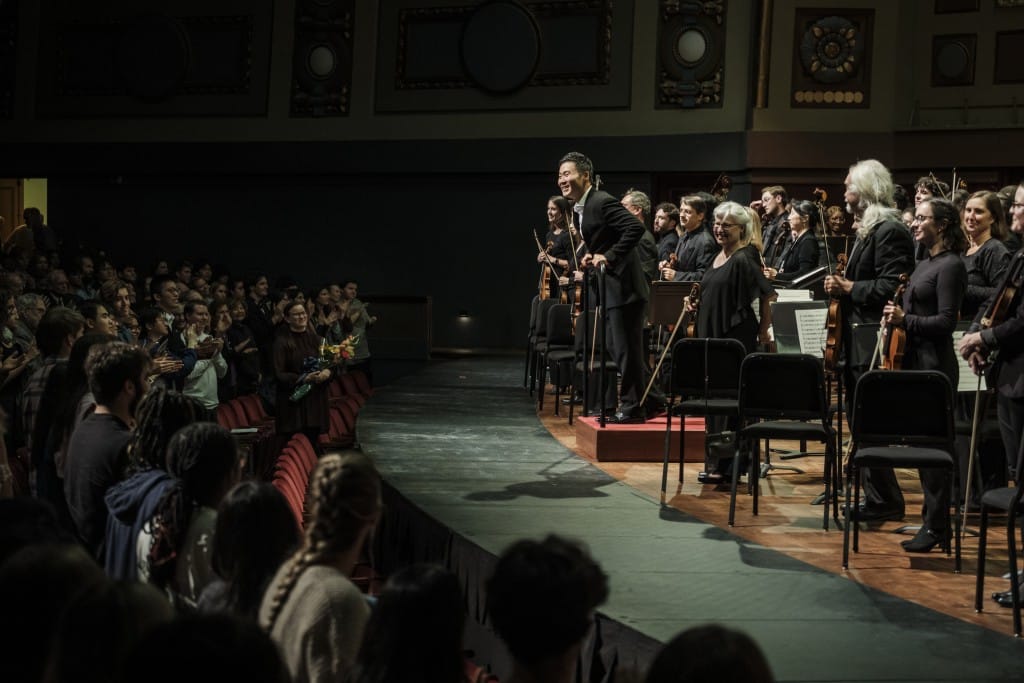 The following image shows an onstage orchestra, led by a conductor, standing to receive applause from an audience, which is also shown standing.