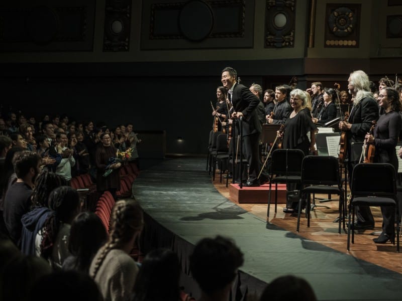 The following image shows an onstage orchestra, led by a conductor, standing to receive applause from an audience, which is also shown standing.